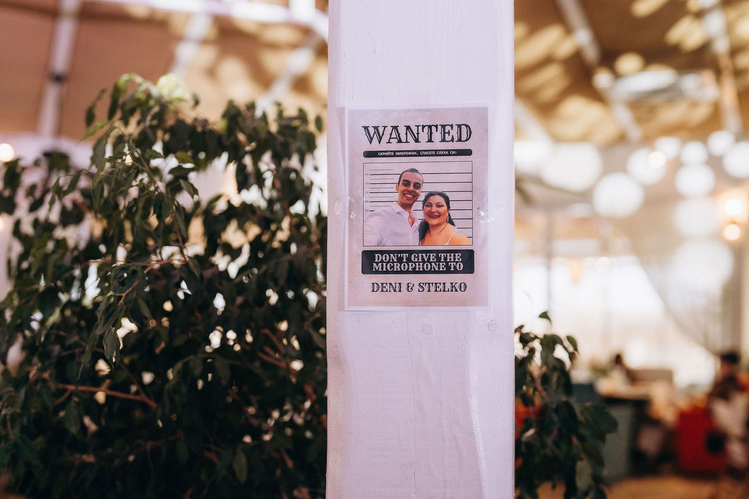 Wanted poster wedding sign featuring couple photo with names Deni and Stelko displayed on white pillar at reception venue.