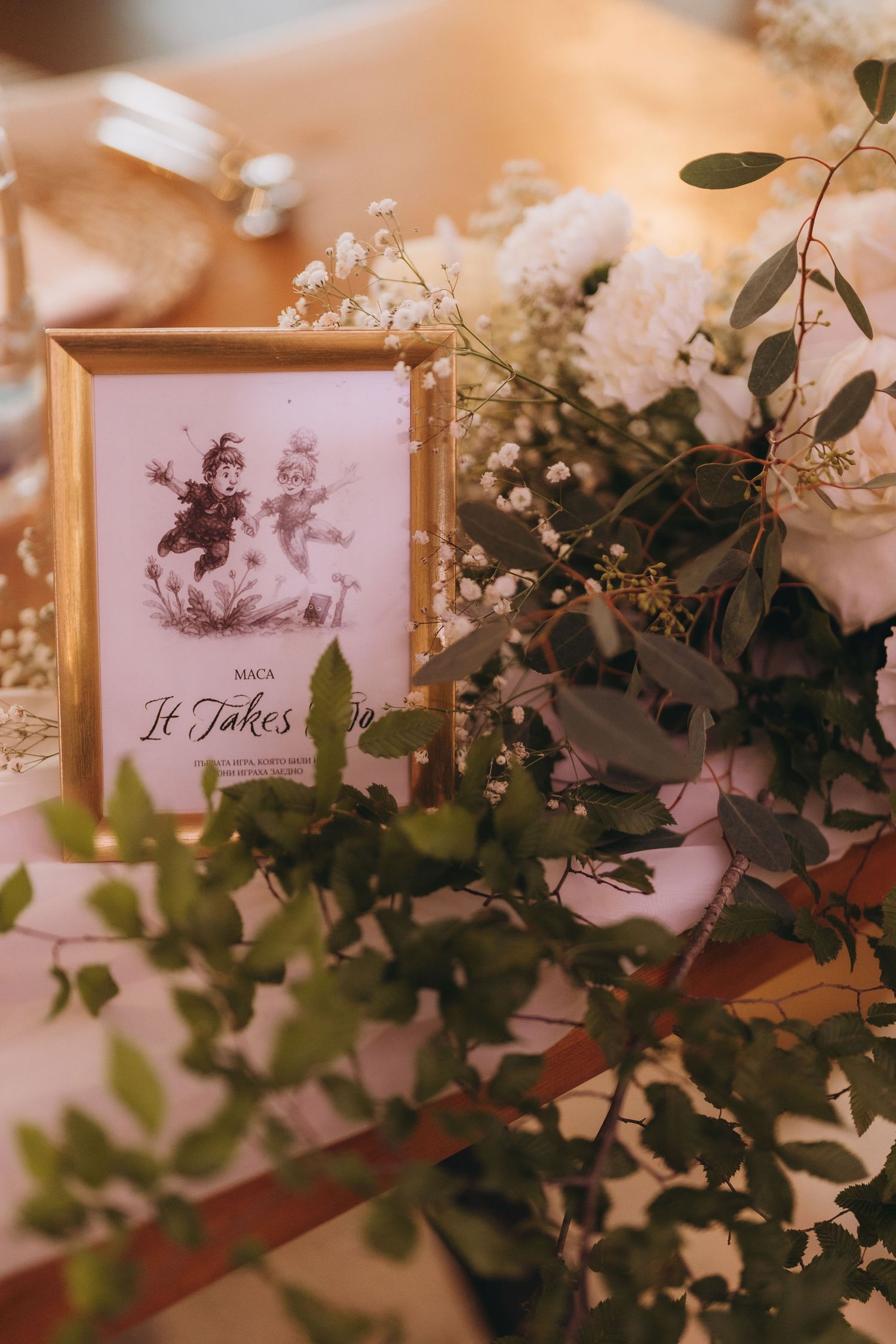 Gold-framed wedding table sign with cherub illustration beside white floral arrangement and eucalyptus greenery.