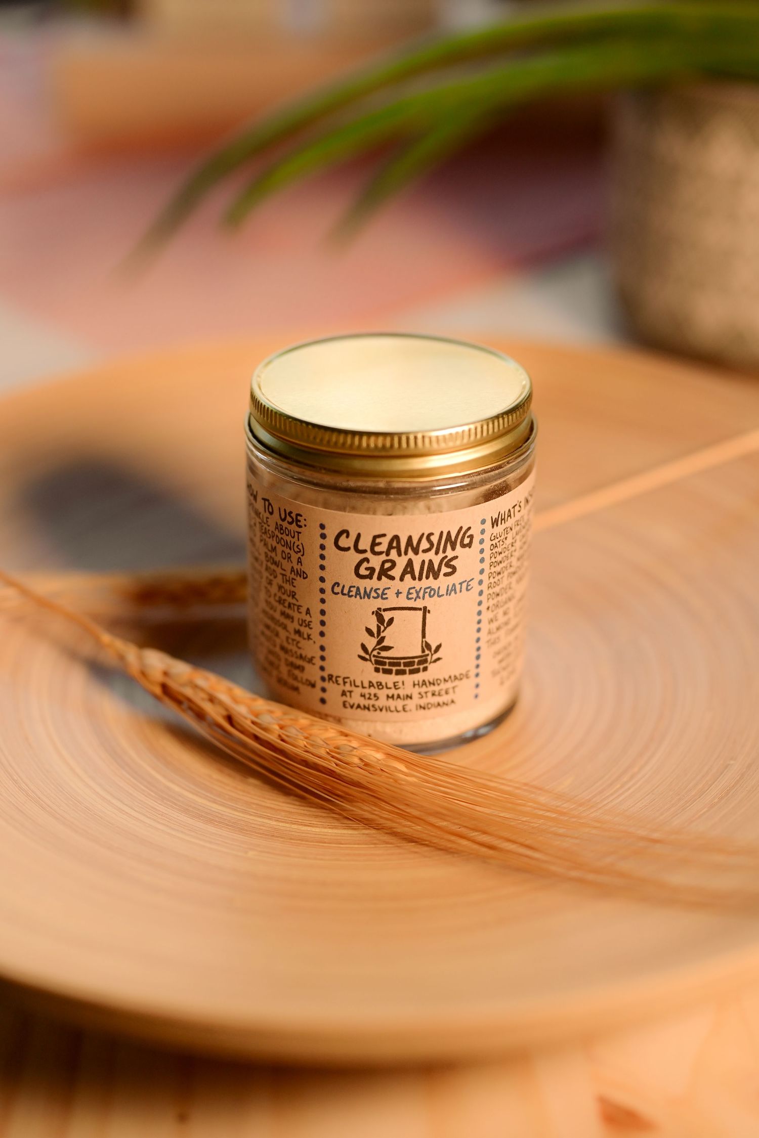 Glass jar of Cleansing Grains facial exfoliant with gold lid, displayed on wooden plate with wheat sprig.