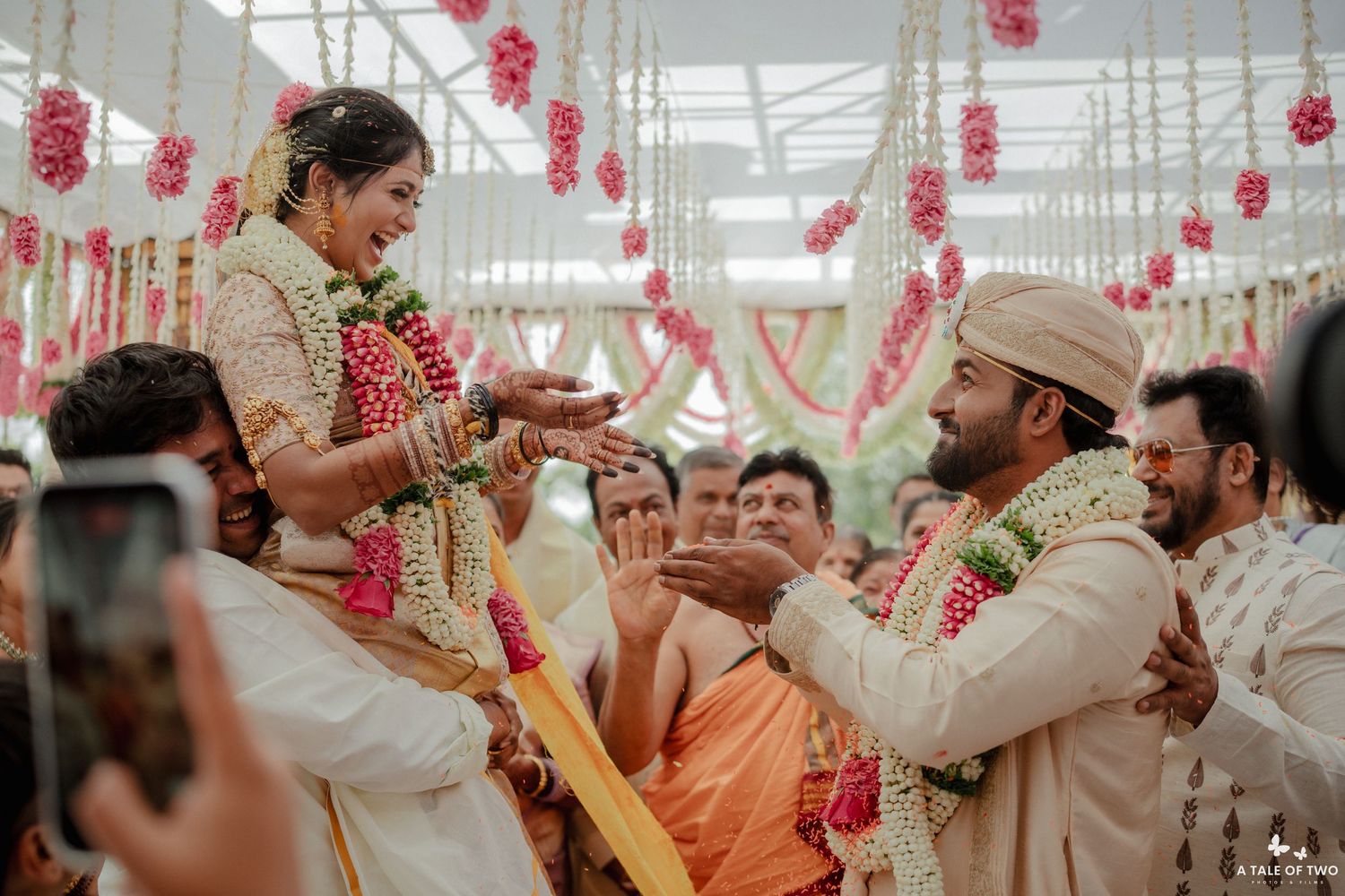 Traditional Indian wedding ceremony with bride lifted by family, exchanging garlands with groom under floral decorations.