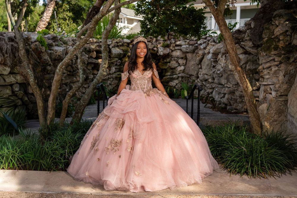 Young woman in pink ball gown quinceañera dress posing outdoors near stone wall and trees.
