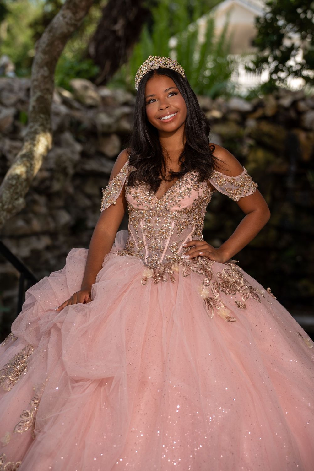 Young woman in pink gold quinceañera ball gown with tiara smiles outdoors near stone wall.