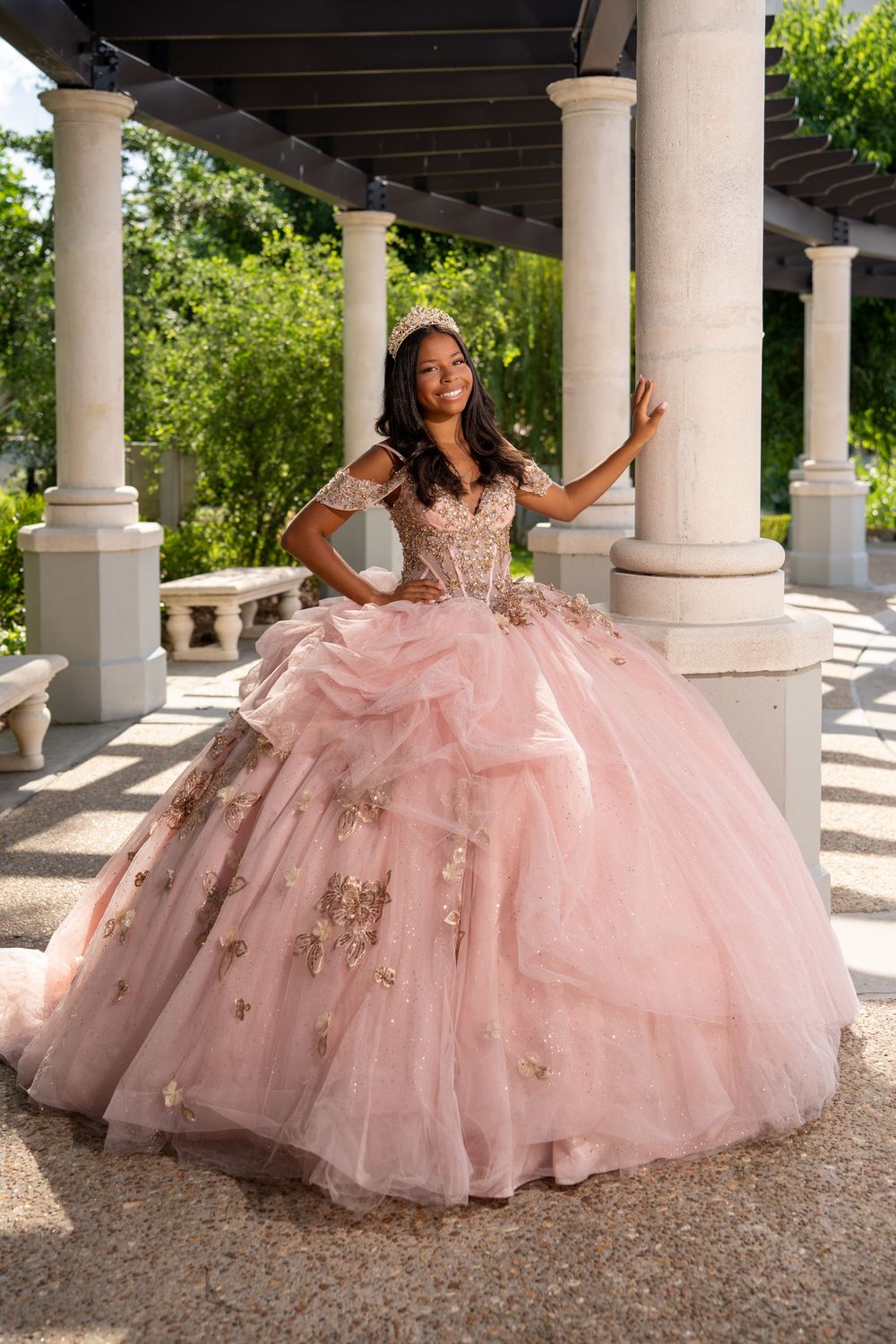 Young woman in a blush pink ball gown with gold floral appliques poses under a pergola surrounded by white columns.