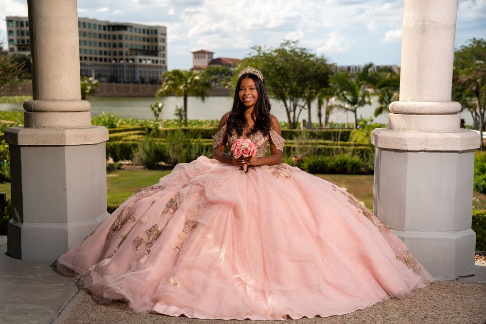 Teen girl in pink quinceañera ball gown holding bouquet under columns with waterfront view.