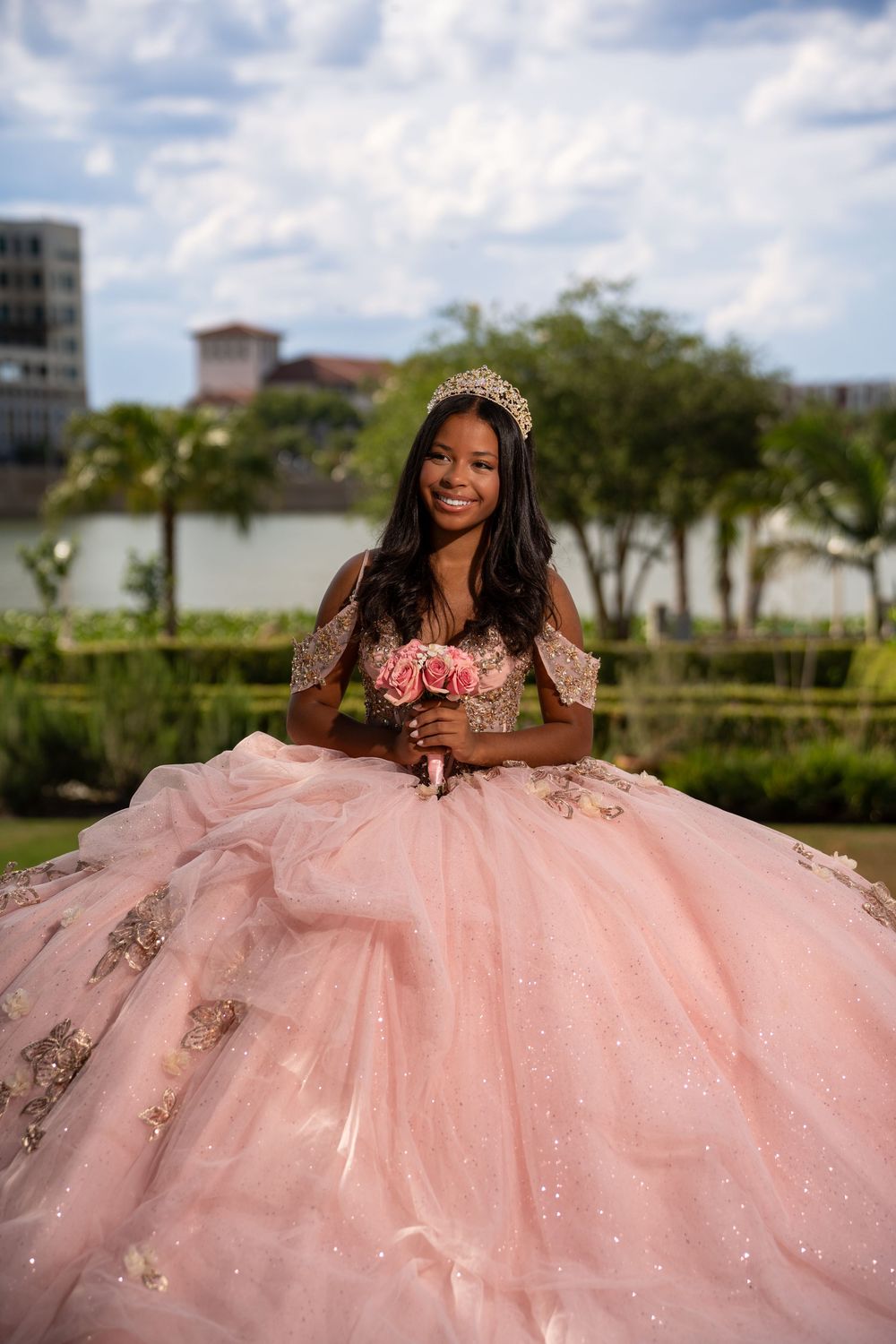 Young woman in pink quinceañera ball gown with tiara holding pink roses bouquet outdoors near lake.