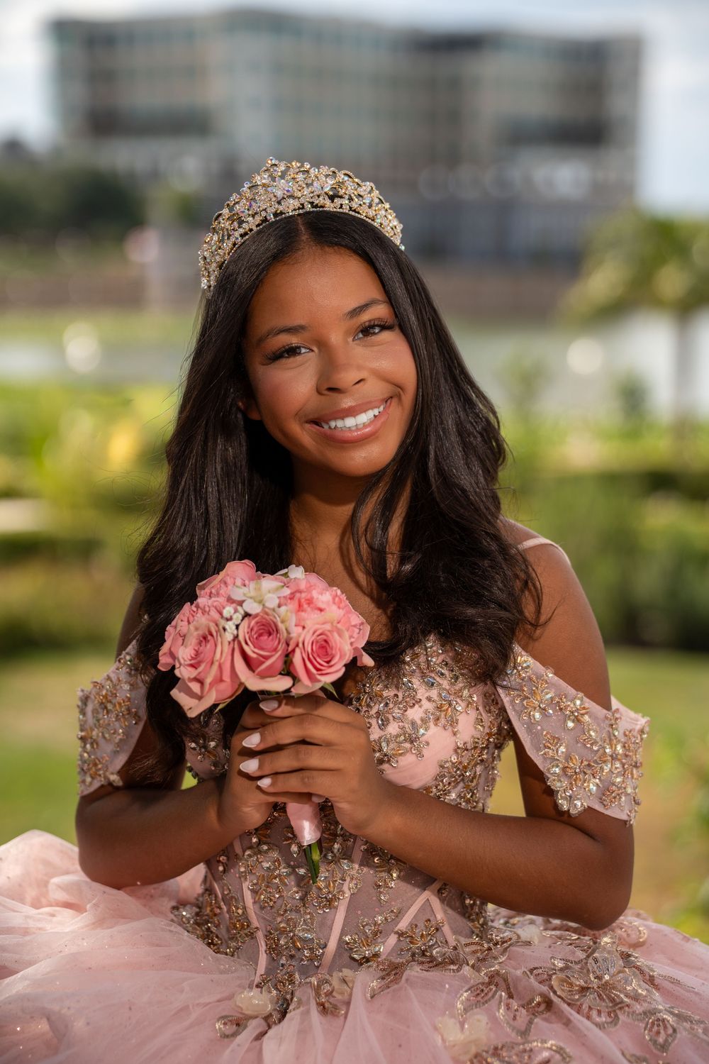 Young woman in pink quinceañera gown with gold tiara holds pink rose bouquet, smiling outdoors.