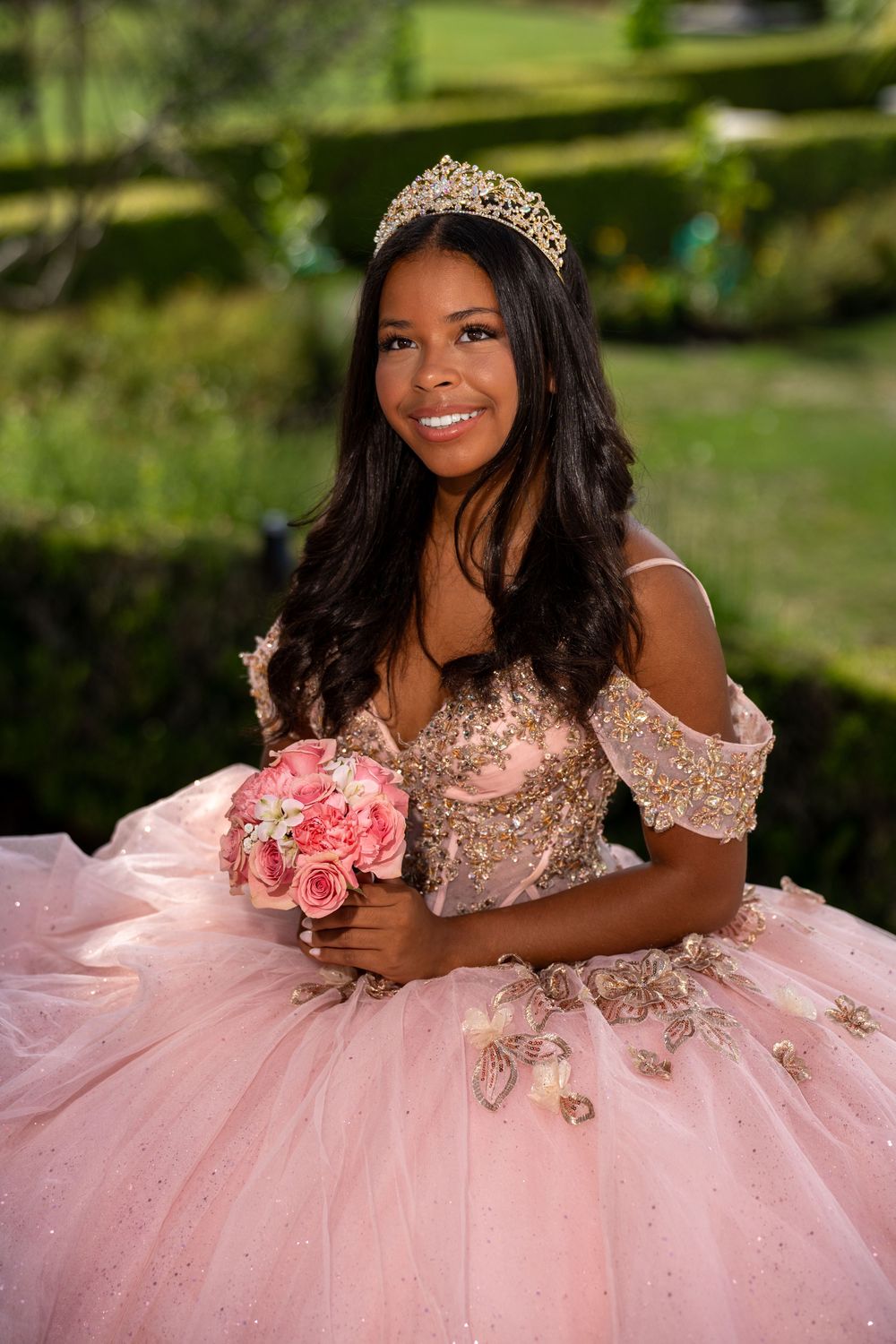 Young woman in pink quinceañera gown with gold tiara holding pink roses bouquet, smiling outdoors.