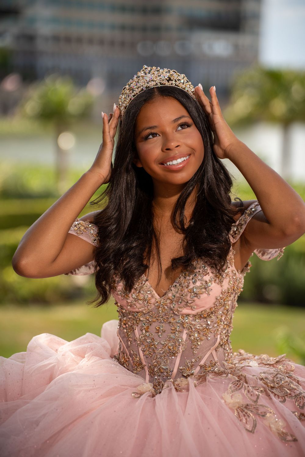 Young woman in pink quinceañera gown and gold tiara smiles joyfully outdoors, adjusting her crown.
