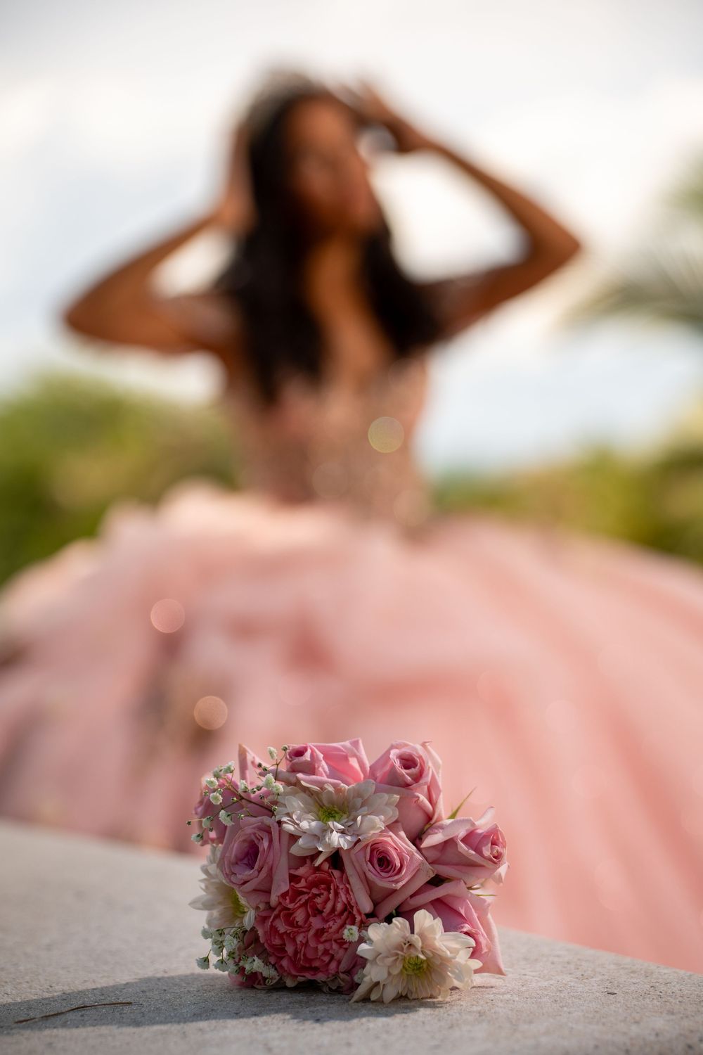 Pink rose bouquet in foreground with blurred quinceañera girl in pink ball gown posing outdoors.