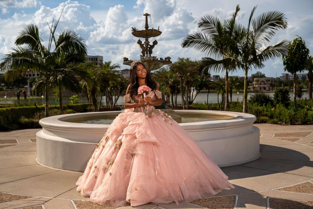 Young woman in pink quinceañera ball gown poses by fountain with palm trees at golden hour.
