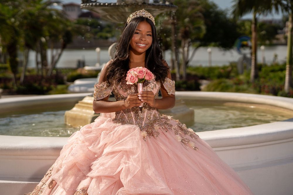 Young girl in pink quinceañera gown with tiara holds bouquet by fountain outdoors.