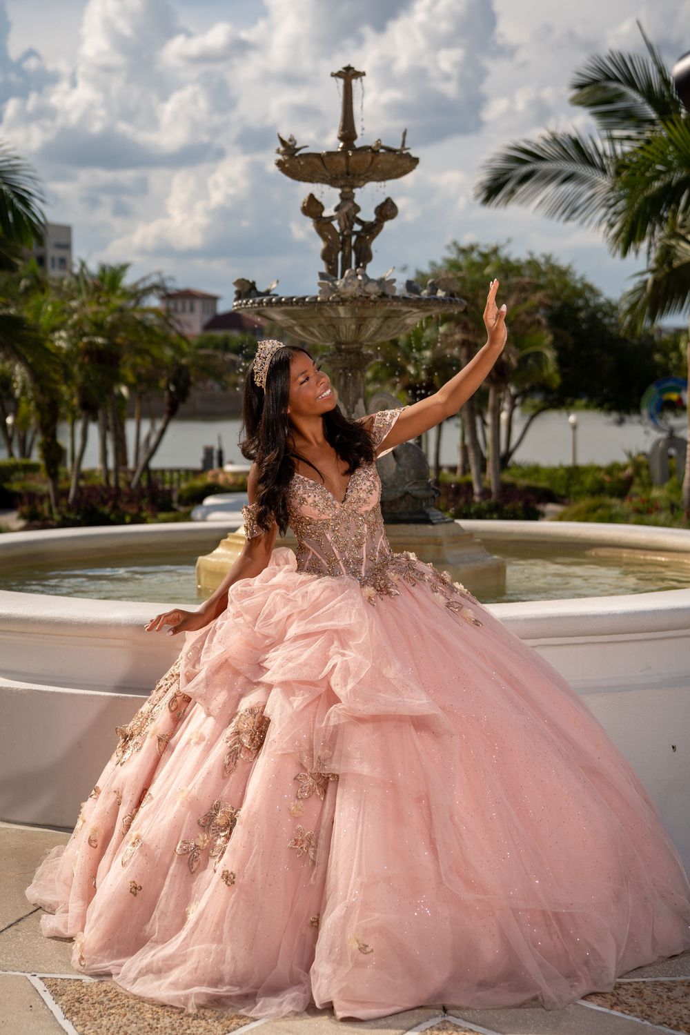 Young woman in pink quinceañera ball gown with gold embroidery poses by ornate fountain outdoors.