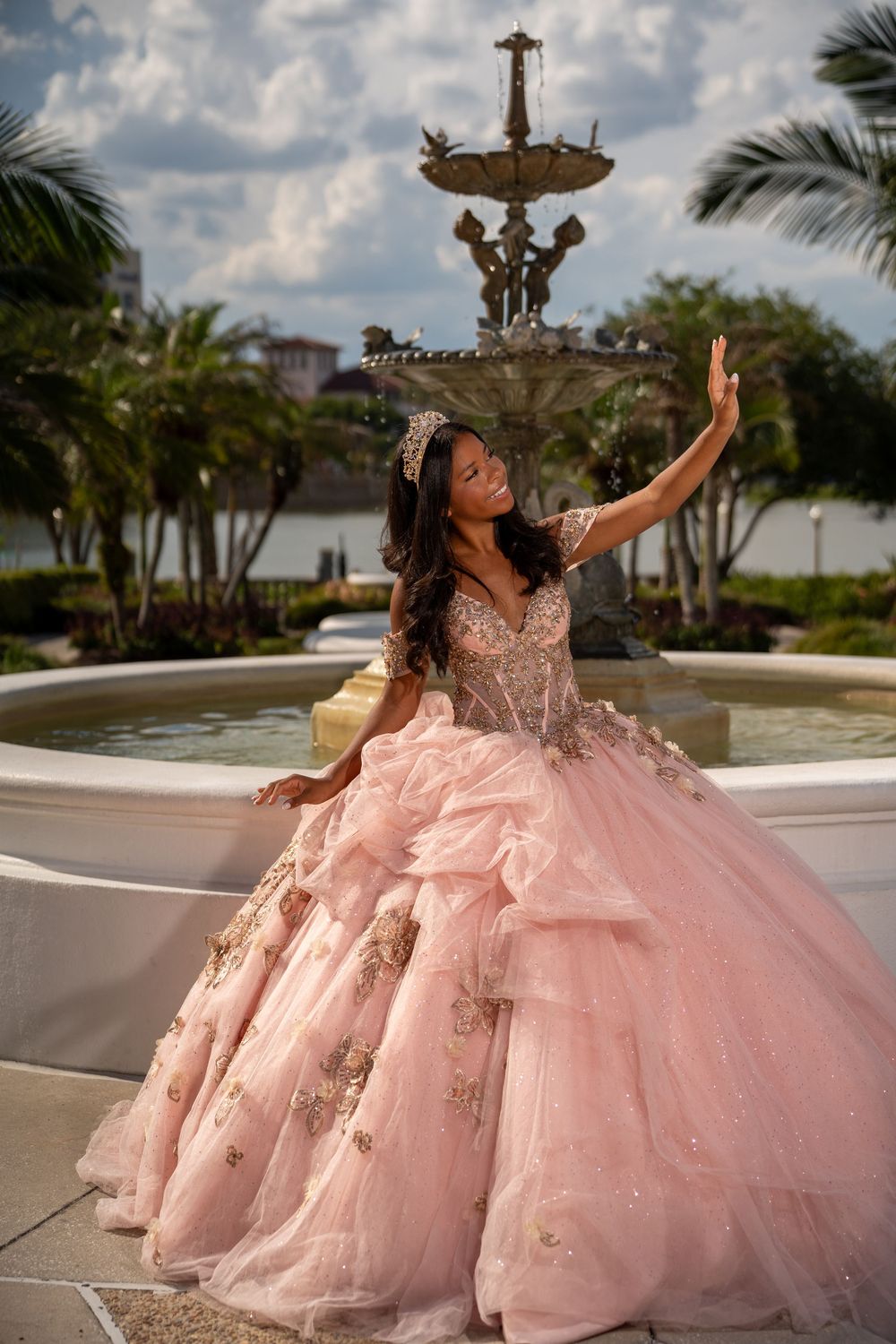 Teenage girl wearing pink tiara and ruffled quinceañera gown waves gracefully near decorative fountain.