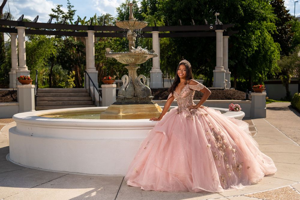 Young woman in a pink floral ball gown poses by a white fountain in a garden for a quinceañera photo.