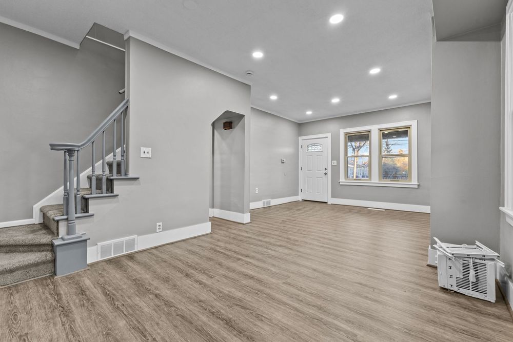 Bright open living room with light gray walls, hardwood floors, recessed lighting, and a staircase.
