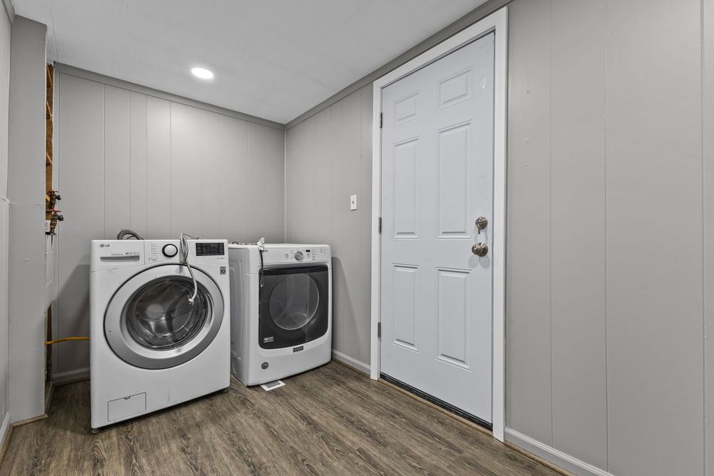 Modern laundry room with side-by-side washer and dryer, gray walls, hardwood floors, and a white door.