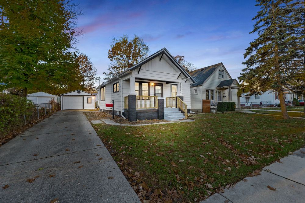 Charming bungalow home with gray siding, covered porch, detached garage, and lush green lawn surrounded by mature trees.