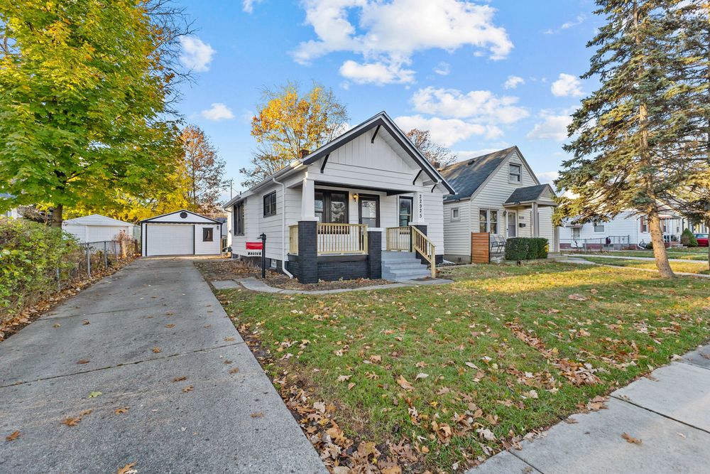 Charming bungalow with gray siding, covered porch, detached garage, and colorful autumn trees on a well-maintained corner lot.