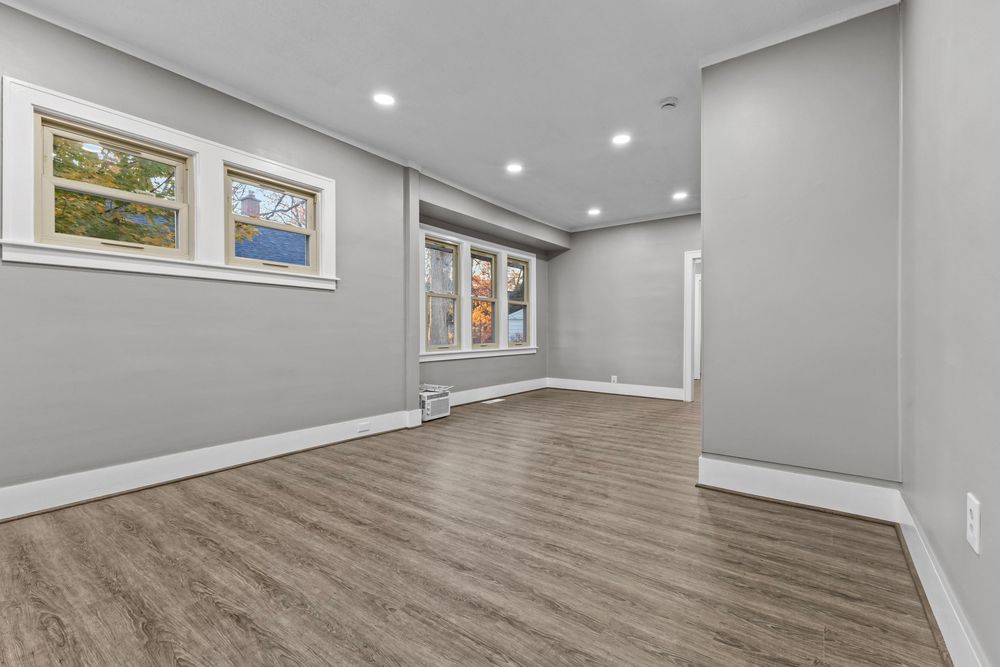 Bright empty room with gray walls, hardwood floors, recessed lighting, and two windows with natural light.