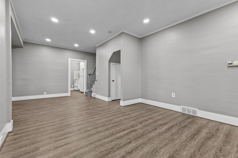 Spacious empty living room with gray walls, hardwood floors, recessed lighting, and an arched doorway leading to a staircase.