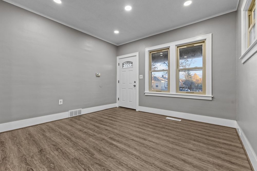Bright empty living room with gray walls, hardwood floors, white front door, and two windows letting in natural light.