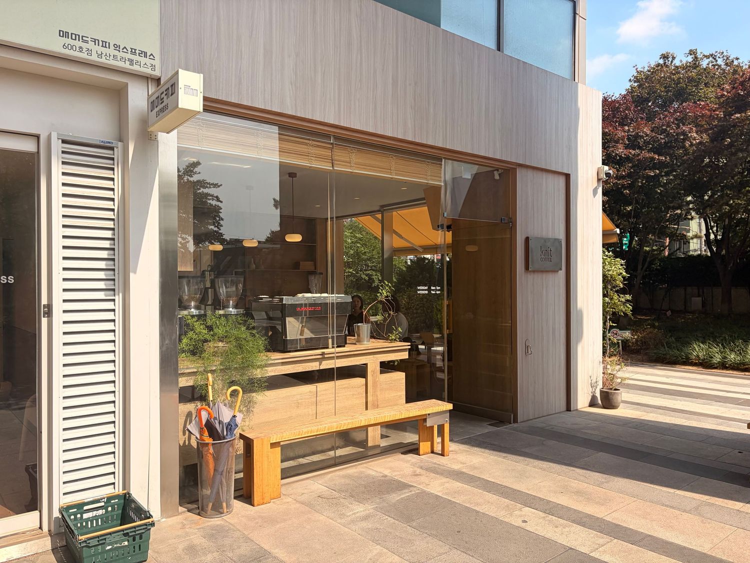 Modern minimalist cafe exterior with wooden bench, glass facade, and lush greenery in an urban setting.