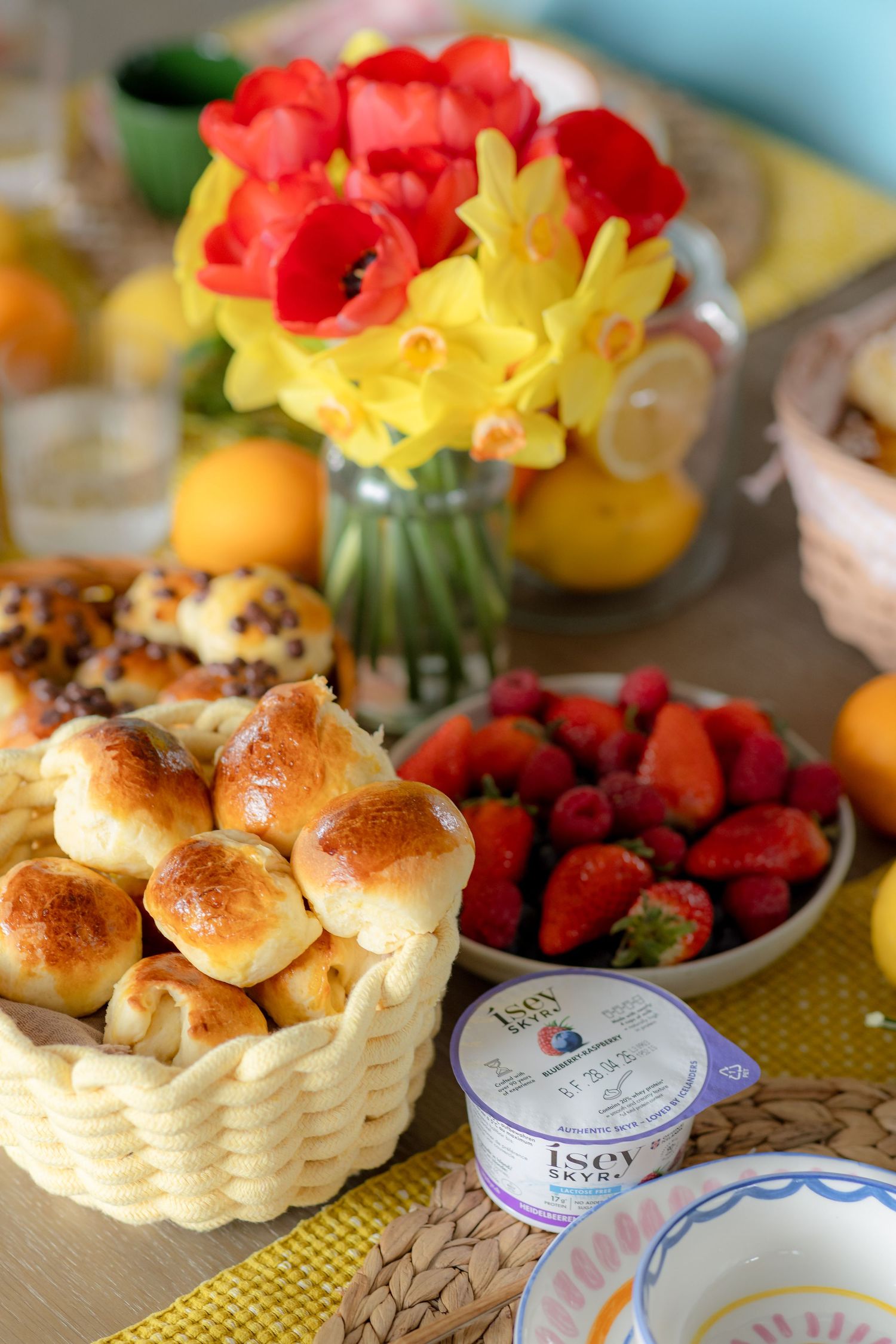 Brunch table with bread rolls, fresh strawberries, Isey Skyr yogurt, and a bouquet of red tulips and yellow daffodils.
