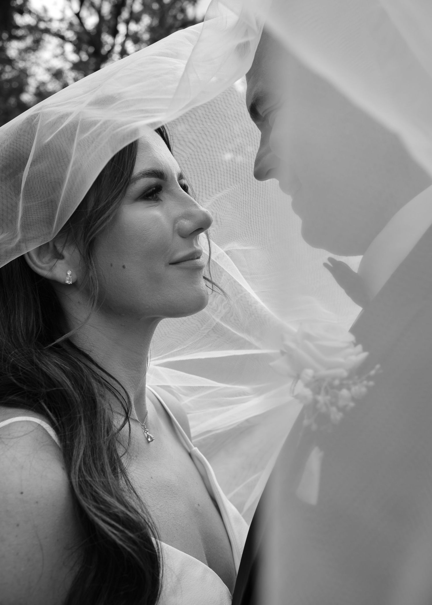 Black and white wedding photo of bride smiling under veil at groom in romantic outdoor portrait.