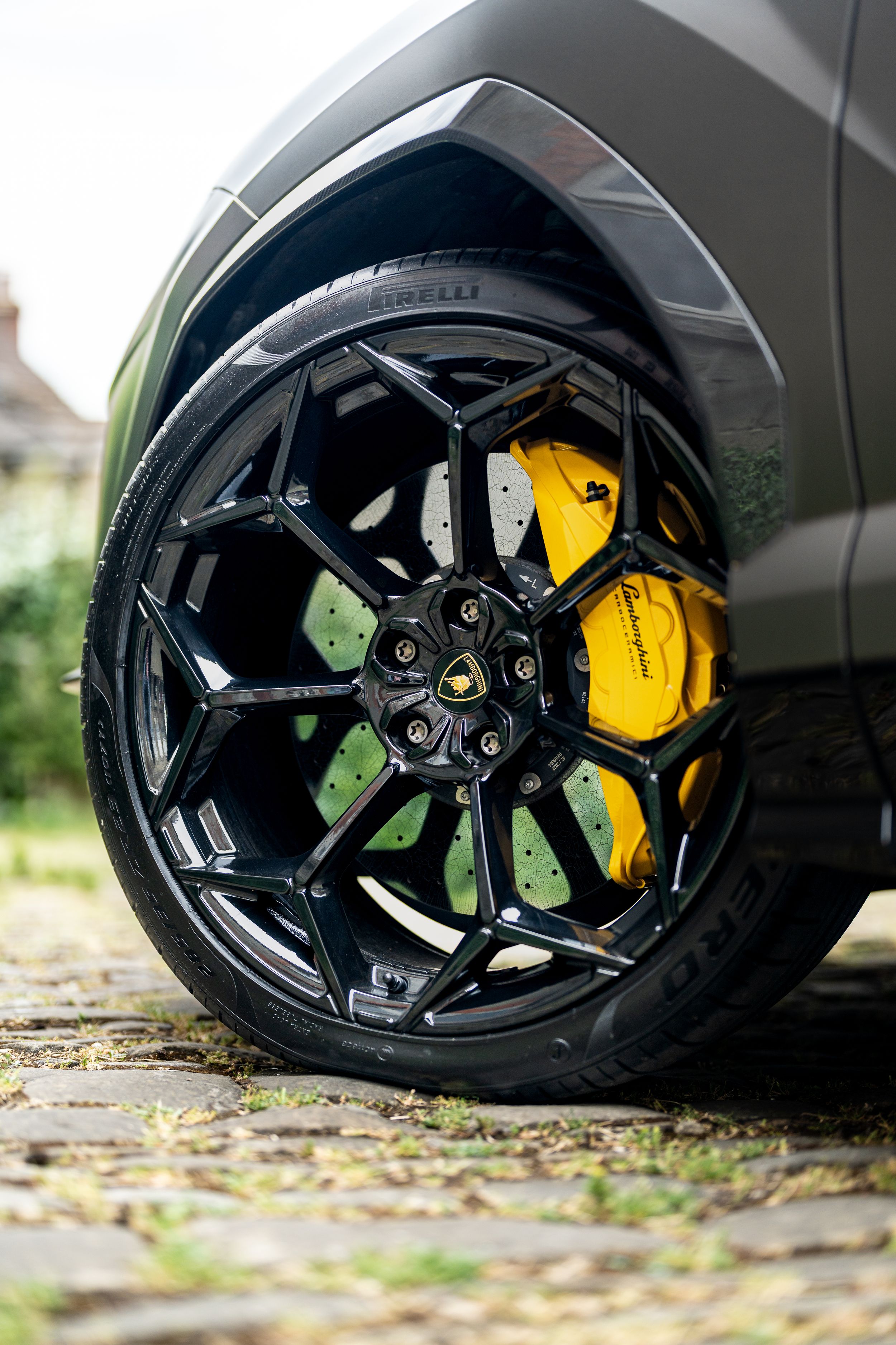 Lamborghini Urus black alloy wheel with yellow brake calipers on a cobblestone surface.