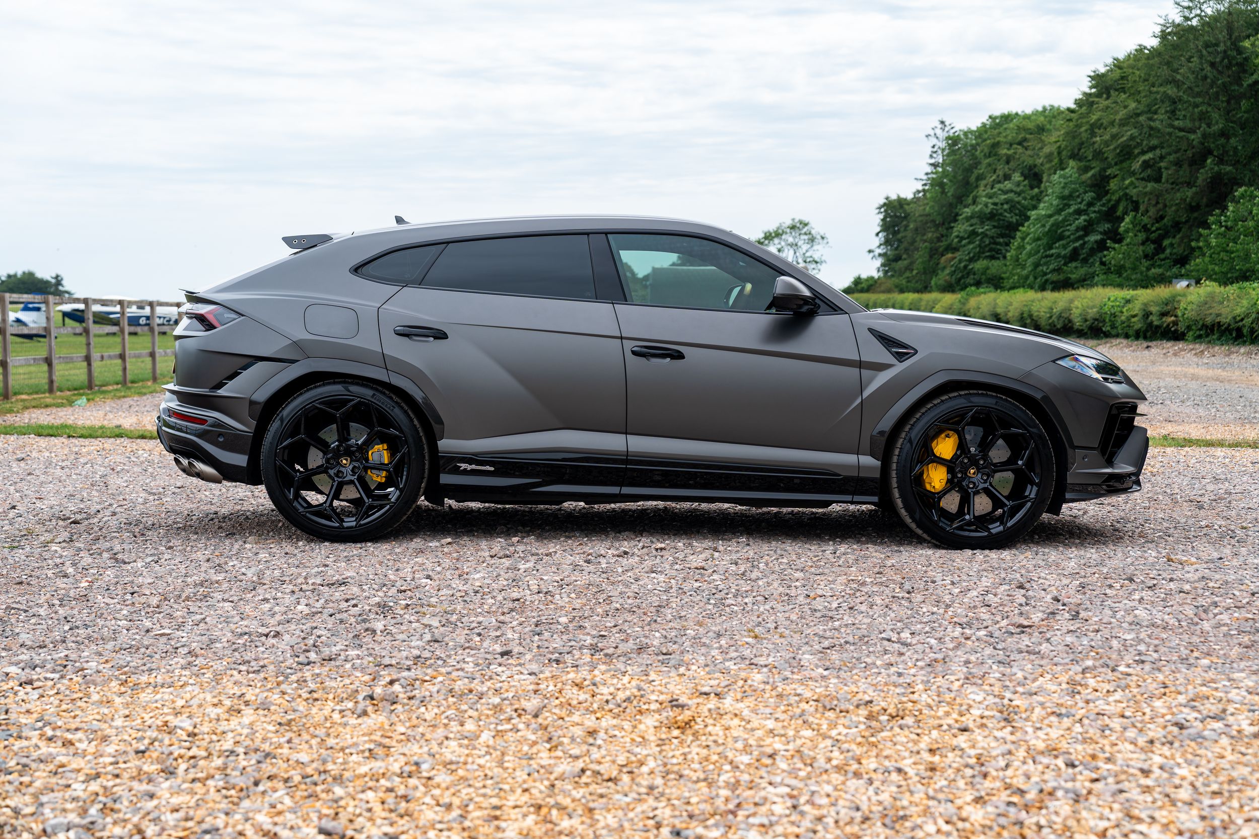 Matte grey Lamborghini Urus SUV with yellow brake calipers parked on gravel driveway surrounded by greenery.