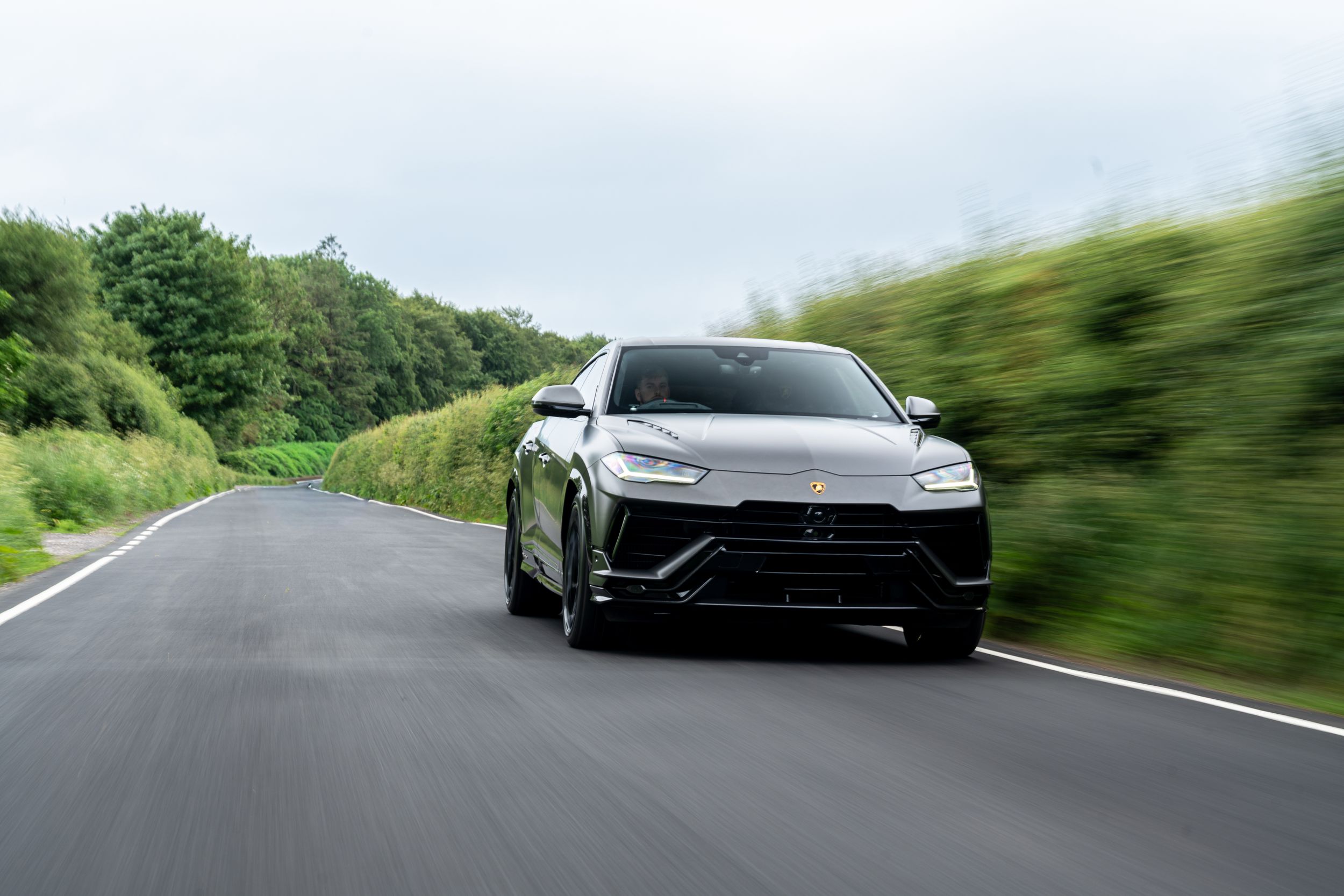 Matte grey Lamborghini Urus Performante driving at speed on a tree-lined country road.