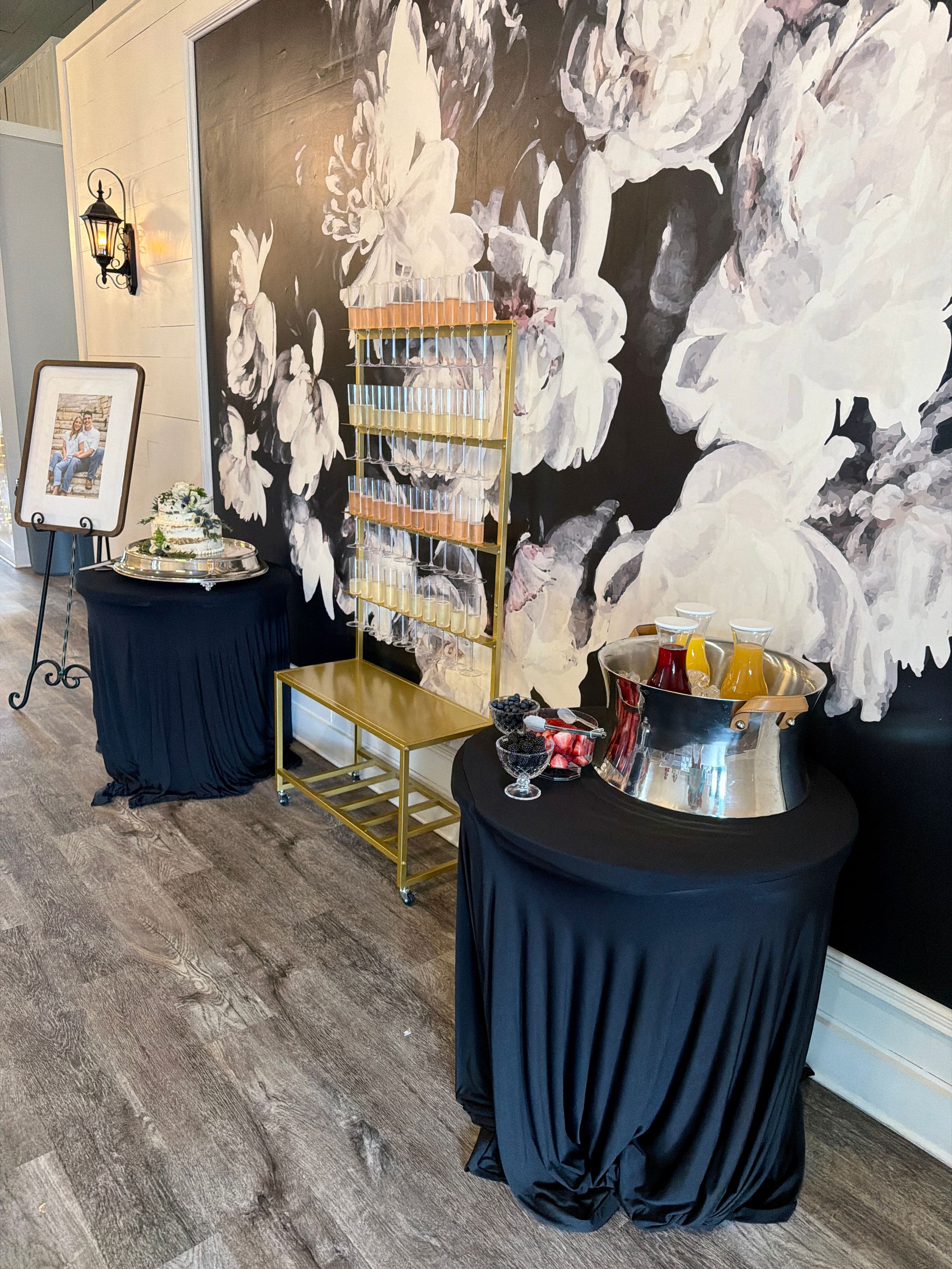 Elegant event space with black floral mural, cocktail tables with black linens, gold shelving, and champagne bucket display.