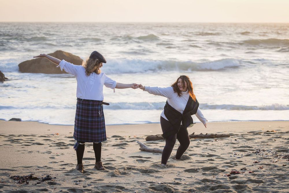 Two women dancing and holding hands on a sandy beach at sunset wearing skirts and white blouses.