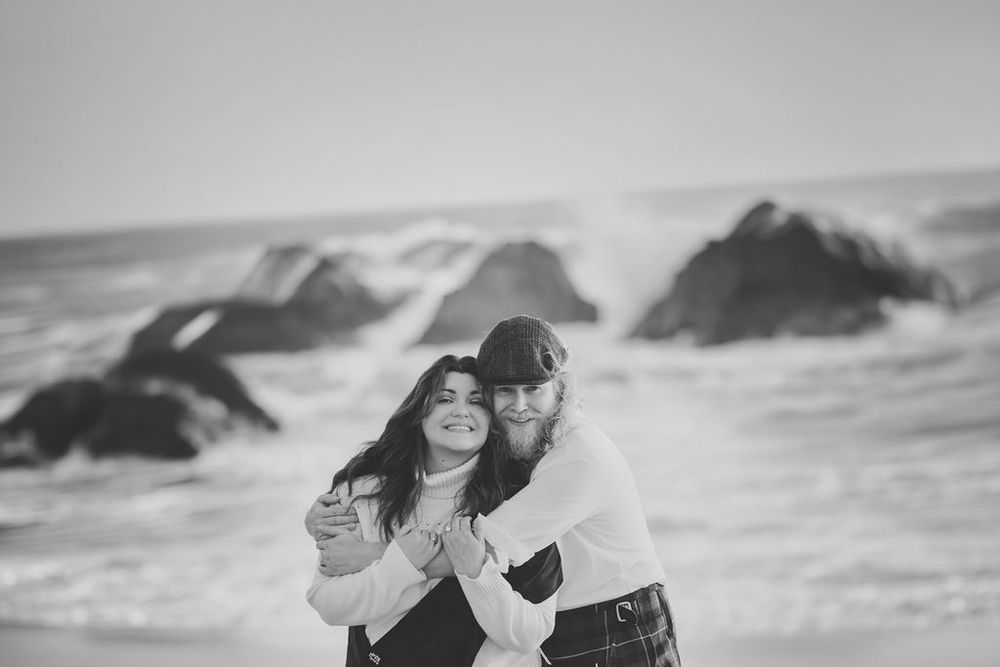 Black and white photo of couple hugging on beach with dramatic ocean waves behind them.