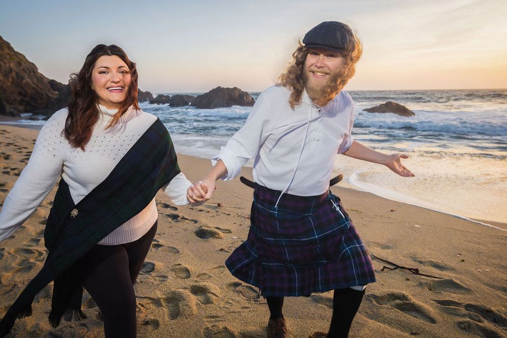 Couple holding hands and dancing playfully on sandy beach at sunset, woman in plaid kilt skirt.