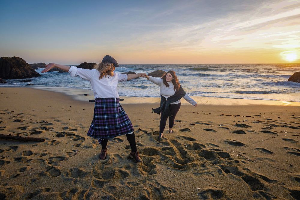 Two women dancing joyfully on a sandy beach at sunset, one wearing a plaid kilt.