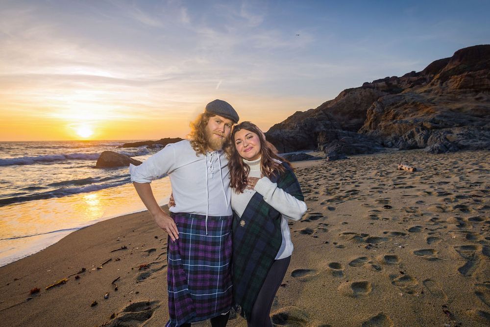 Couple in Scottish kilts embracing on sandy beach at golden sunset with rocky coastline.