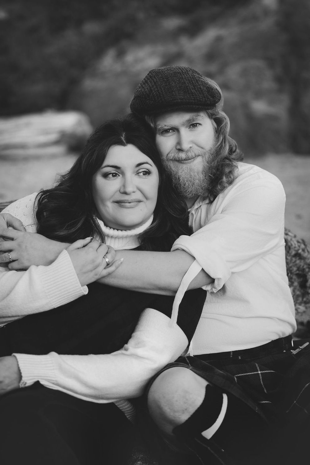 Black and white photo of bearded man in flat cap embracing dark-haired woman outdoors.