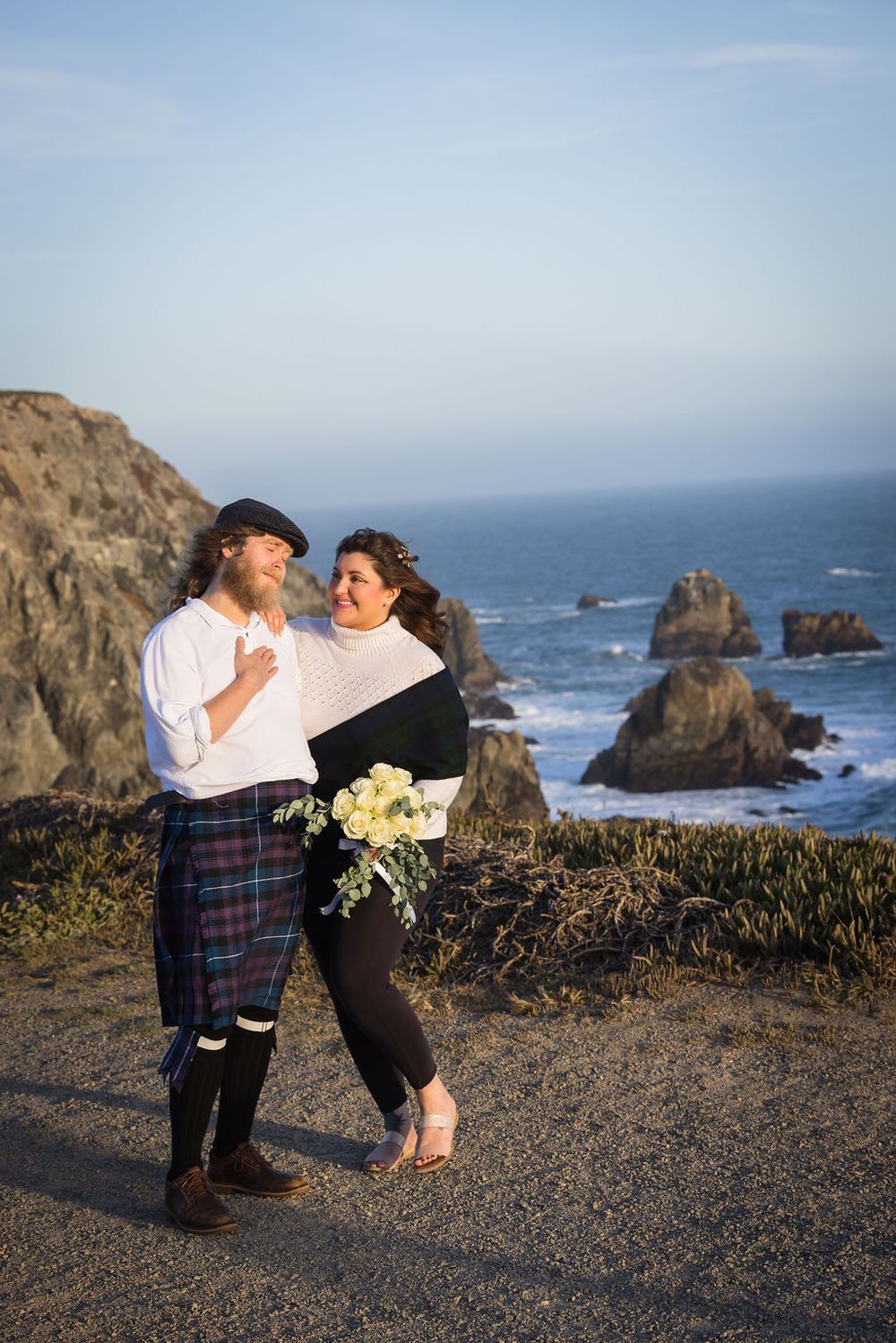 Couple laughing together on coastal overlook, man in tartan kilt holding bouquet of white flowers.