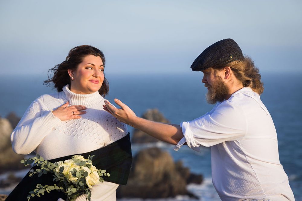Bearded man in flat cap adjusting white sweater of dark-haired woman holding bouquet with ocean backdrop.