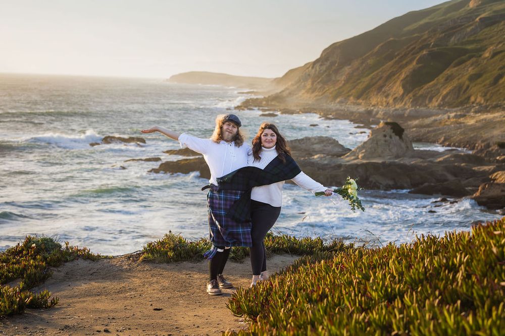 Two women pose playfully on a rocky coastal cliff at golden hour, one carrying the other with ocean waves behind them.