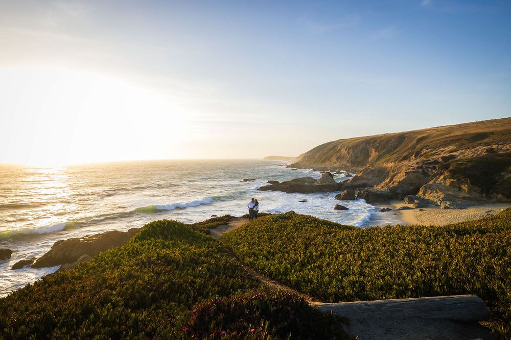 Person sitting on a grassy coastal cliff overlooking a rocky beach at golden sunset with rolling hills in background.