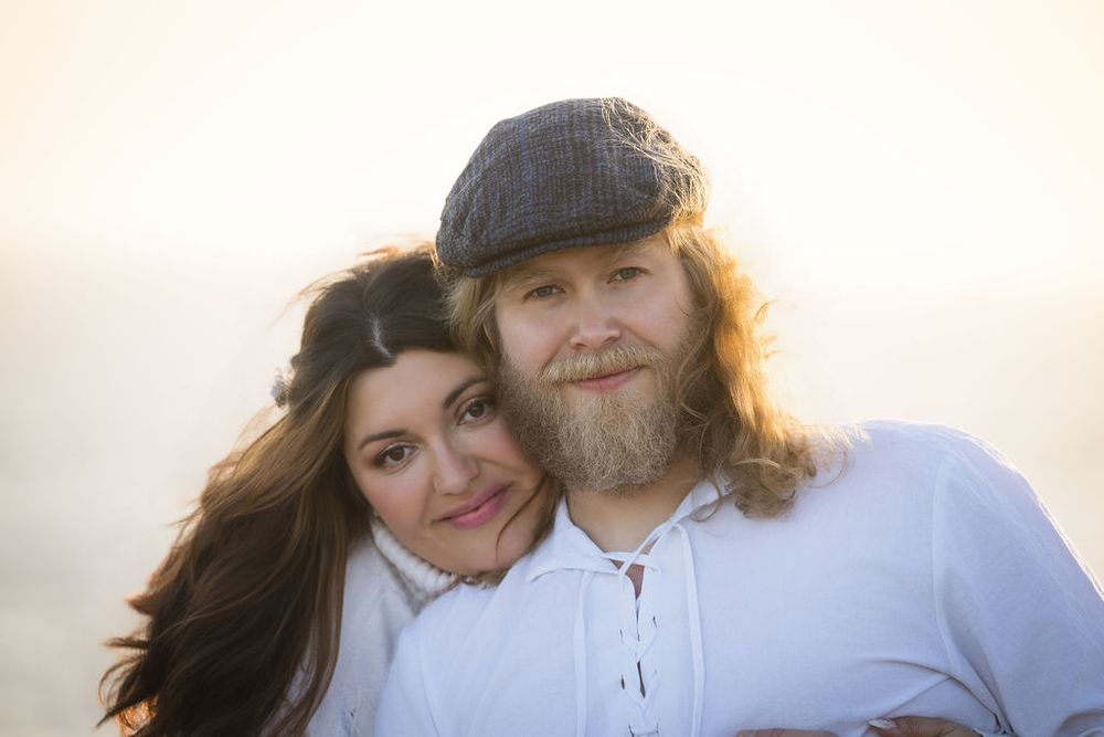 A smiling couple poses closely together in warm golden backlight, man with beard wearing a gray flat cap embracing woman.