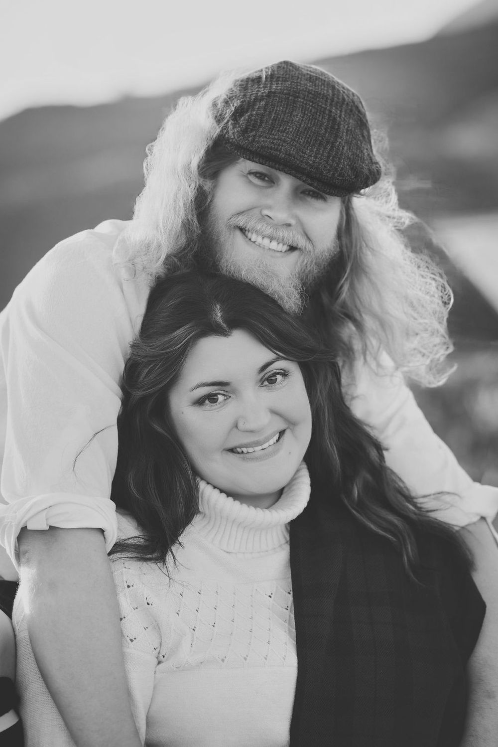 Black and white portrait of smiling couple, man in flat cap with long curly hair resting chin on womans head.
