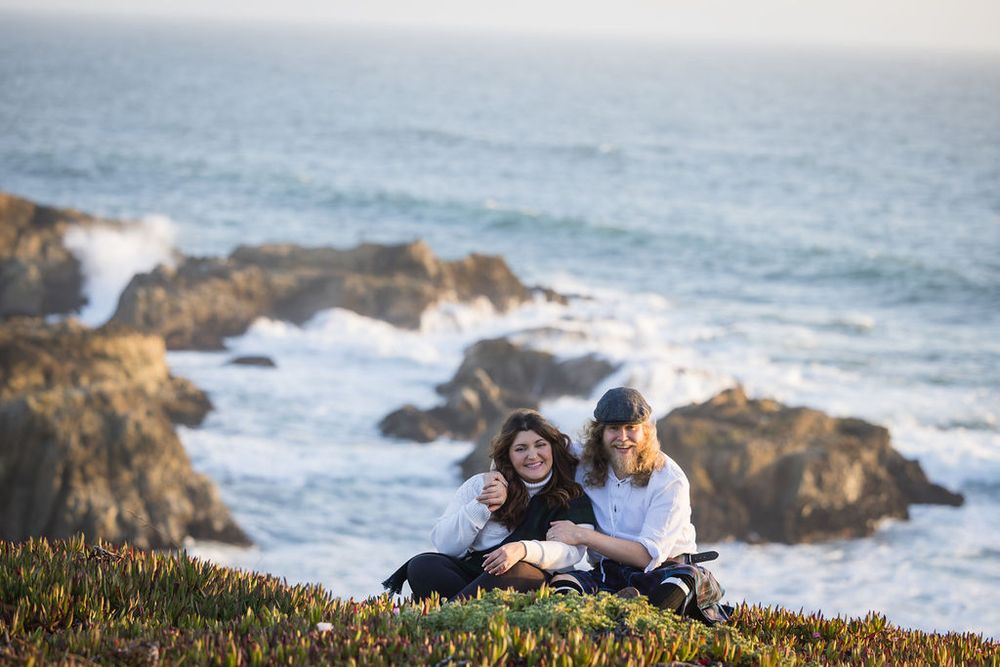 Couple sitting together on coastal cliff overlooking rocky shoreline and ocean waves at golden hour.