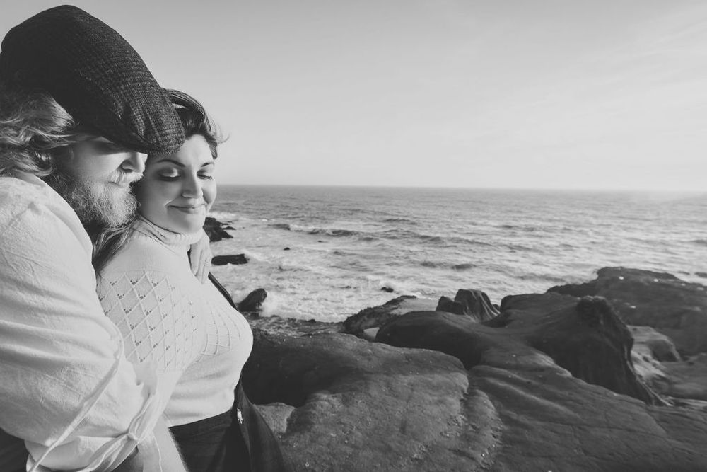 Two women share a tender moment on rocky coastal cliffs overlooking ocean waves in a black and white photo.