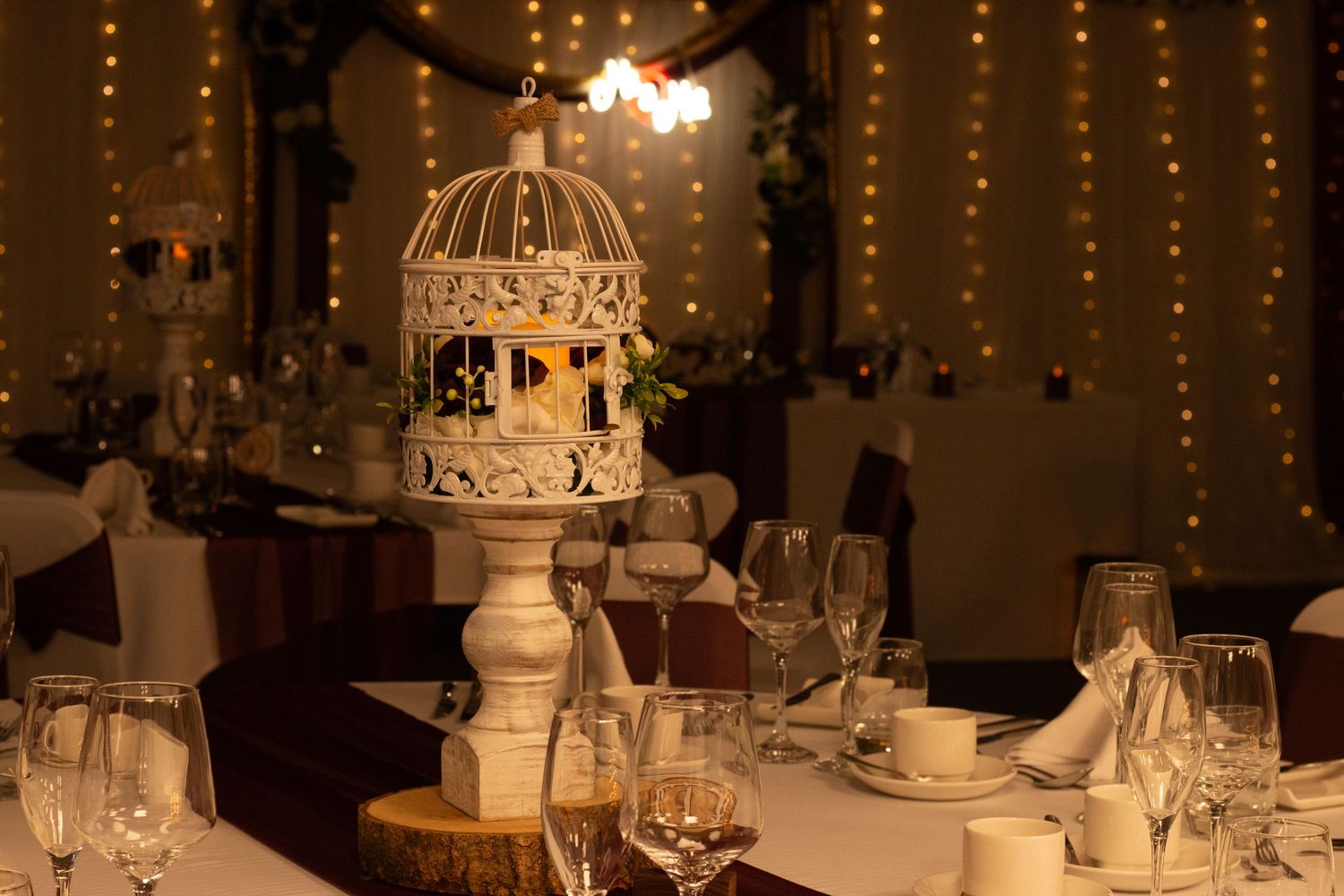 Elegant wedding reception table with white birdcage centerpiece, fairy lights, crystal glassware, and soft romantic ambiance.