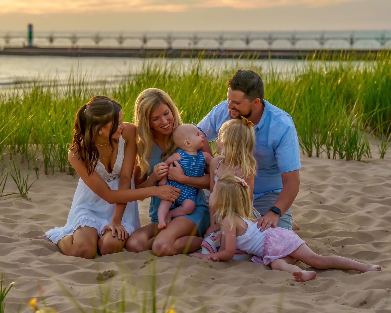 Family of five enjoying a golden sunset beach session on sandy dunes near a pier, with parents and children smiling together.