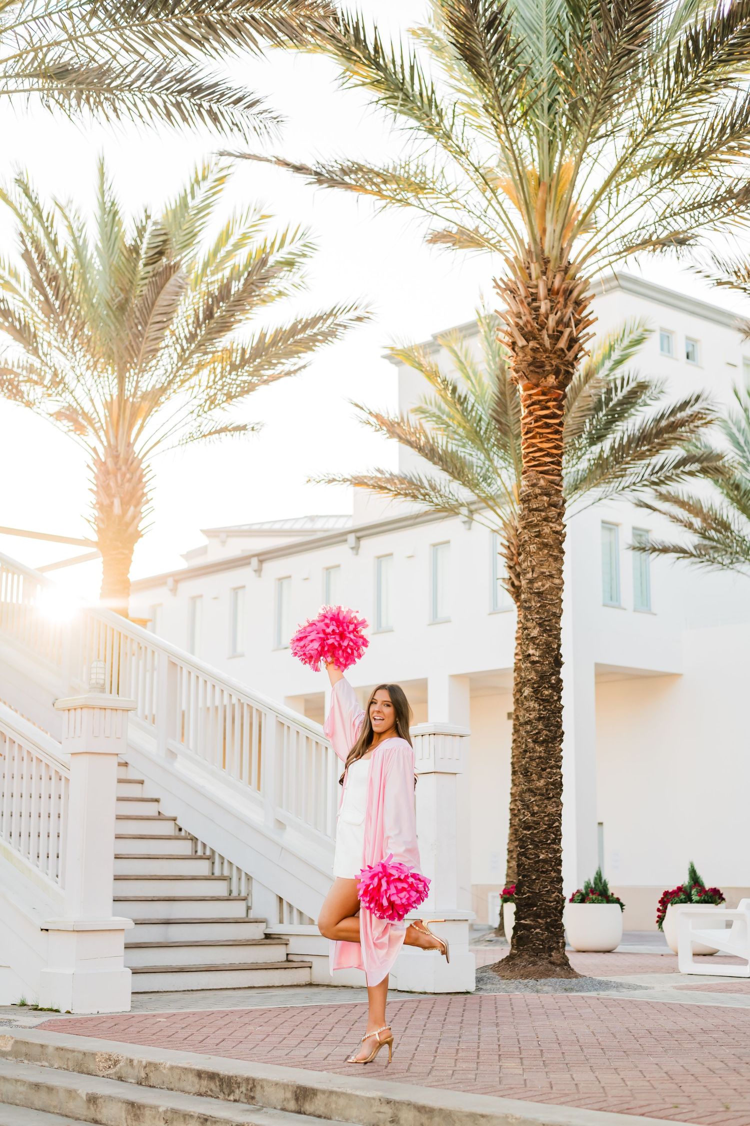 Young woman in pink graduation gown holding pink pom-poms near palm trees and white building staircase.