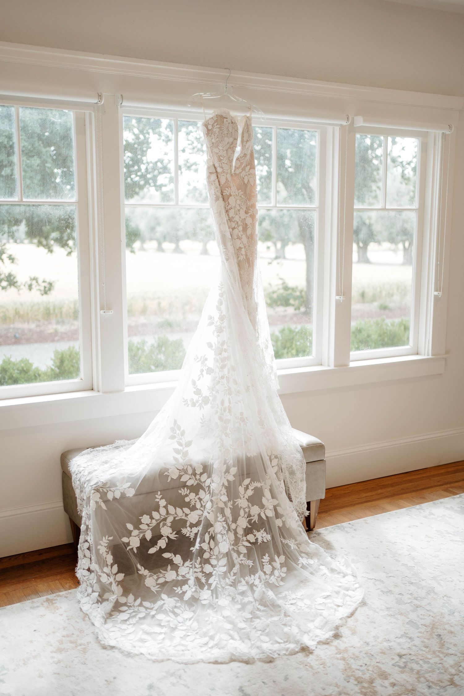 Elegant lace wedding dress with floral appliqués hanging in a bright window, with a long train draped over a bench.